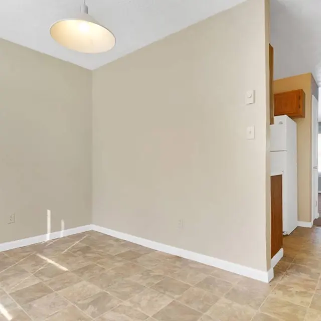 An empty room with beige walls and a light fixture hanging from the ceiling, featuring tiled flooring. A doorway leads to another room and a refrigerator is visible in the background.