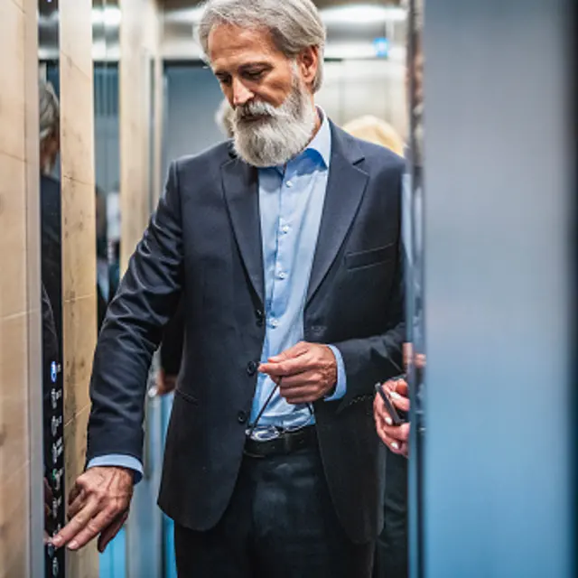 A mature businessman with a gray beard in a suit stands inside an elevator, looking at the control panel while pressing a button.
