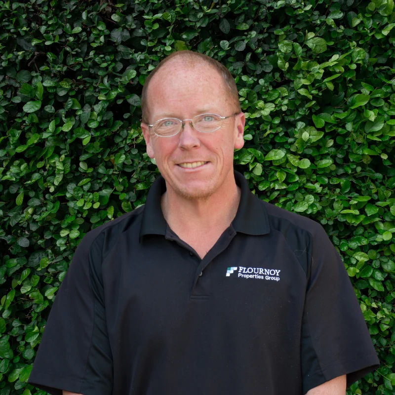 A man with glasses wearing a black shirt with a logo stands in front of a green leafy background, smiling at the camera.