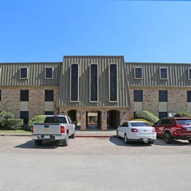 Front view of a multi-story brick building with a green metal roof, surrounded by well-manicured shrubs and parked cars.