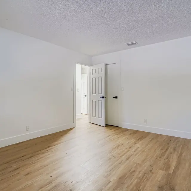 A spacious, empty room with light wooden flooring and white walls. There are two doors visible, one slightly ajar leading to another room or bathroom.