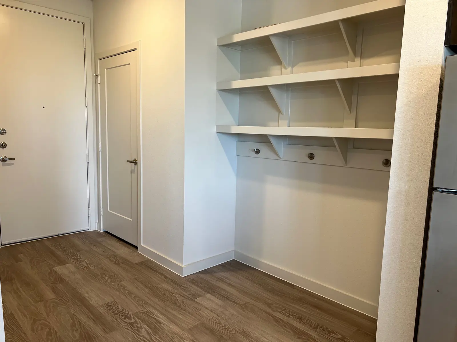 An entryway featuring a door on the left and a closed closet door. To the right, there are white shelves mounted on the wall, and the floor has light wood flooring.