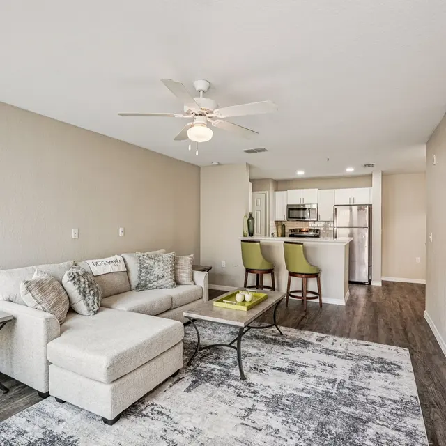 A modern living room featuring a sectional sofa, a glass coffee table, a decorative vase, and a ceiling fan. The room has light-colored walls and a textured rug, leading to an open kitchen area with bar seating.