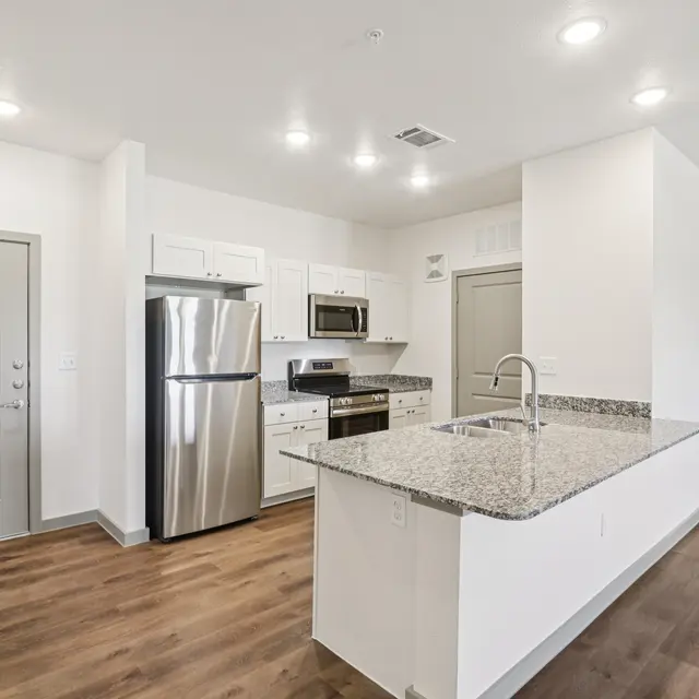 A modern kitchen featuring stainless steel appliances including a refrigerator and microwave, a large granite countertop, and white cabinets. The flooring is a warm wooden texture and the walls are painted white, creating a bright and airy space.