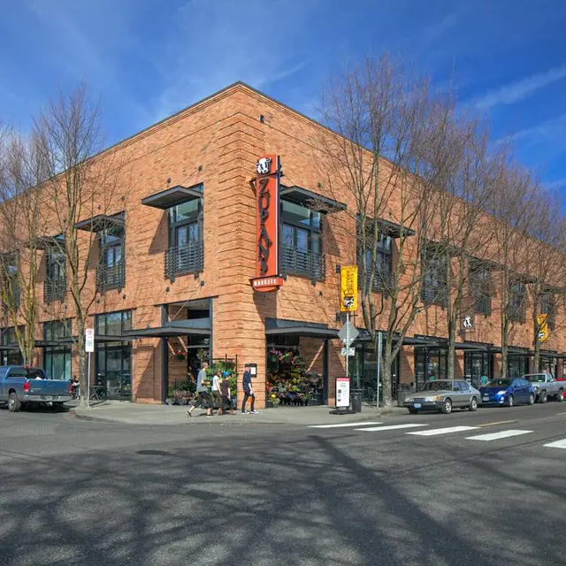 A modern brick building on a street corner with trees and cars parked nearby, under a blue sky with some clouds.