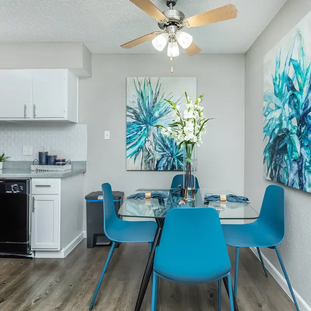 A modern dining area with a glass table and blue chairs, featuring abstract floral artwork on the walls and a ceiling fan above.