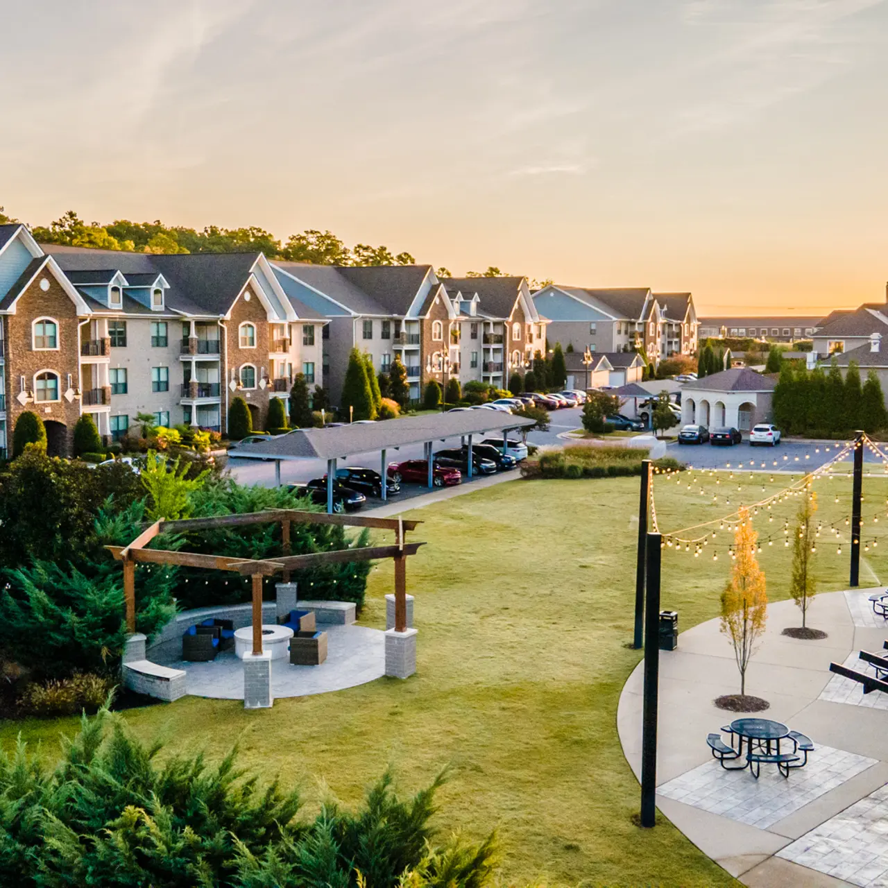 Photo of the green space at The Pointe Brodie Creek in Little Rock, Arkansas. There is a fire pit, three grilling stations, and three picnic tables. Covered parking, a mailhouse, and several community buildings are in the background.
