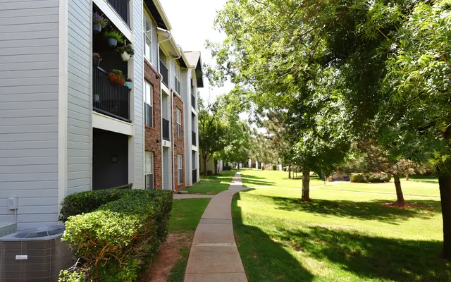 A path lined with greenery between apartment buildings, with trees and well-maintained landscaping on a sunny day.