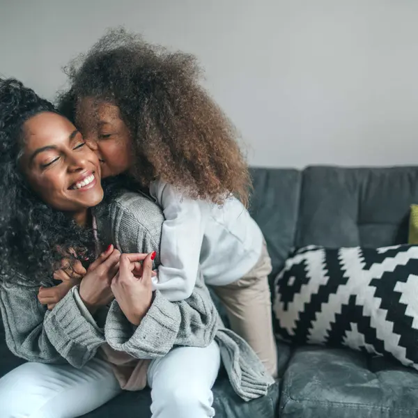 A joyful moment between a mother and child, with the child playfully kissing the mother's cheek while they sit on a couch.