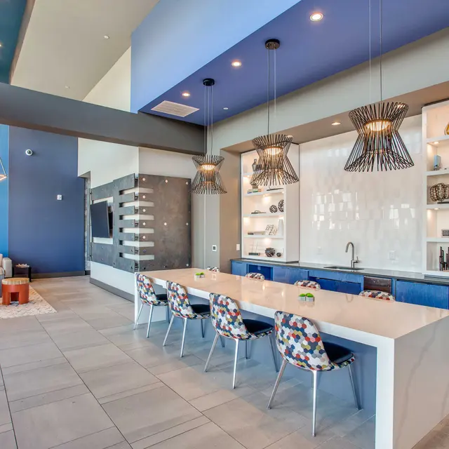A modern and stylish living space featuring a large white table with colorful chairs, a blue kitchen area with shelves displaying various decorative items, and vibrant pendant lights overhead. Soft seating area with a couch and orange accents is visible in the background.