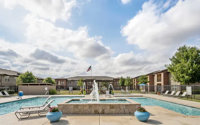 A clear pool surrounded by lounge chairs and a fountain, with trees and buildings in the background under a partly cloudy sky.