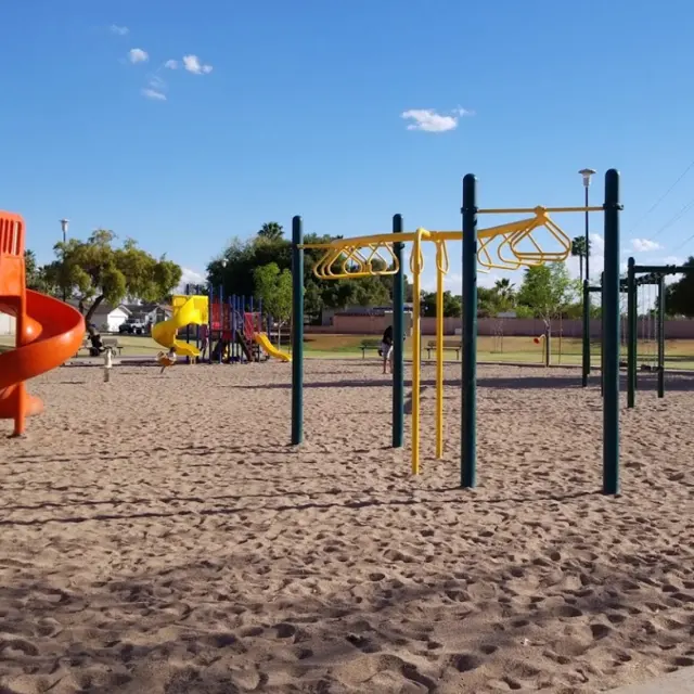 A playground featuring various play structures such as slides, monkey bars, and climbing equipment situated on sandy ground. The sky is clear blue with a few clouds.