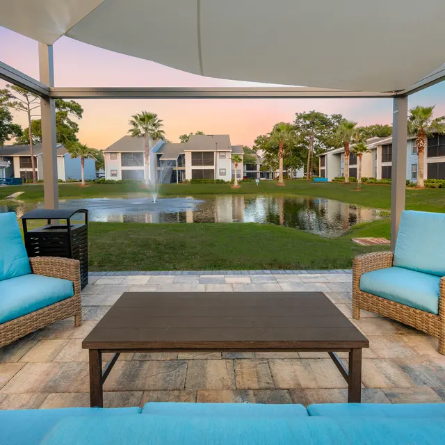 Covered patio seating with cushioned seats and coffee table, by a pond with a fountain in the center.