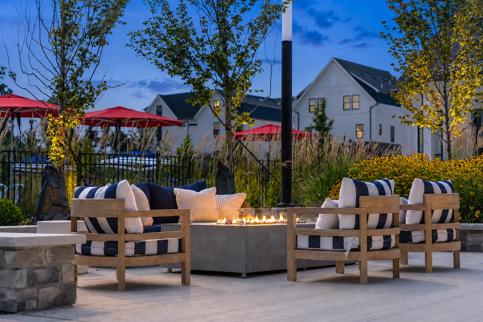 Outdoor lounge area with comfortable seating and a fire pit surrounded by trees and flowers at dusk.