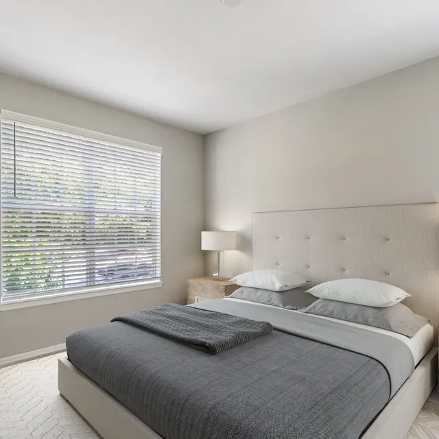 A cozy modern bedroom featuring a neatly made bed with gray bedding, two bedside tables with lamps, and a large window with blinds letting in natural light.