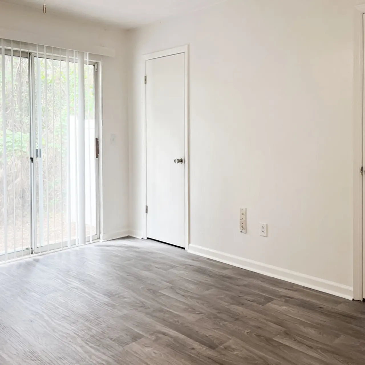 A spacious, empty room with gray laminate flooring, featuring a large sliding glass door that provides access to an outdoor area, and a white door leading to another space. The walls are painted white, creating a bright and airy atmosphere.