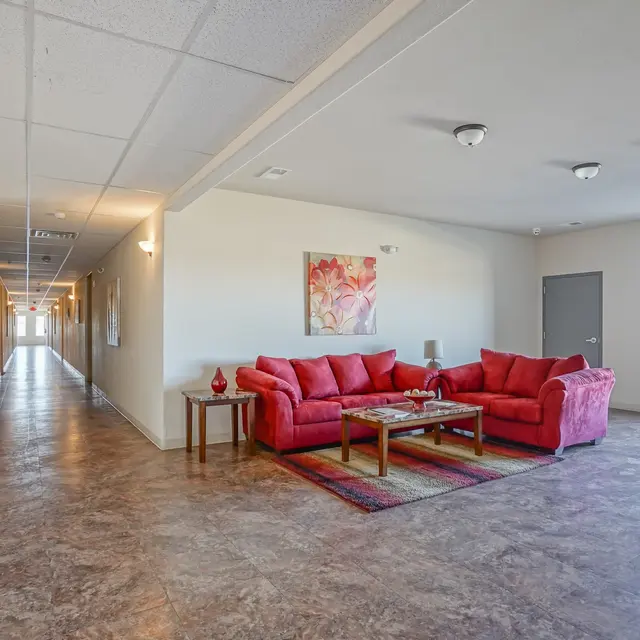 A welcoming lobby area featuring a red sectional sofa, a wooden coffee table, and a small area rug on tiled flooring, with a long hallway visible in the background.