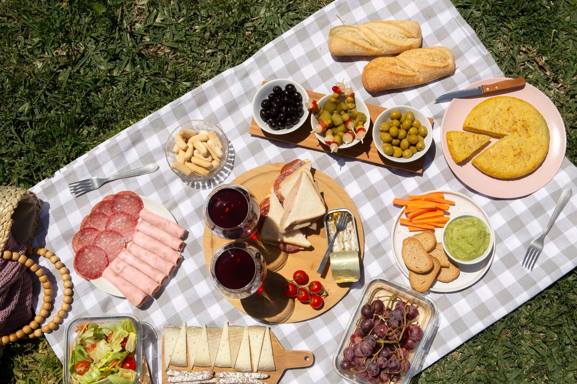 Picnic Spread with Various Delicacies A picnic spread featuring various foods on a checkered blanket, including bread rolls, meats, cheese, olives, a frittata, fruits, and drinks.