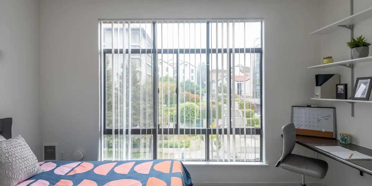 A modern bedroom featuring a bed with a colorful comforter, a large window with vertical blinds, and a desk arranged with a chair and decorative items.