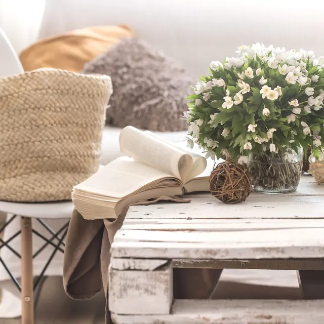A cozy living space featuring a white chair, a woven basket, a vase of flowers, an open book, and decorative spheres on a rustic table.