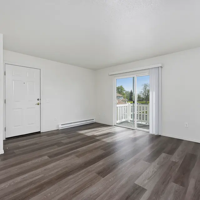 Empty Living Room An empty living room featuring light-colored walls and dark wood-like flooring. There is a front door on the left and a sliding glass door leading to a balcony on the right, with natural light coming in. The room is spacious and uncluttered.