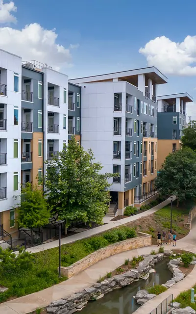 Modern mid-rise exterior of 120 Ninth Street Apartments in San Antonio, showcasing large windows and private balconies, with a landscaped walking path beside the iconic San Antonio River Walk landmark.