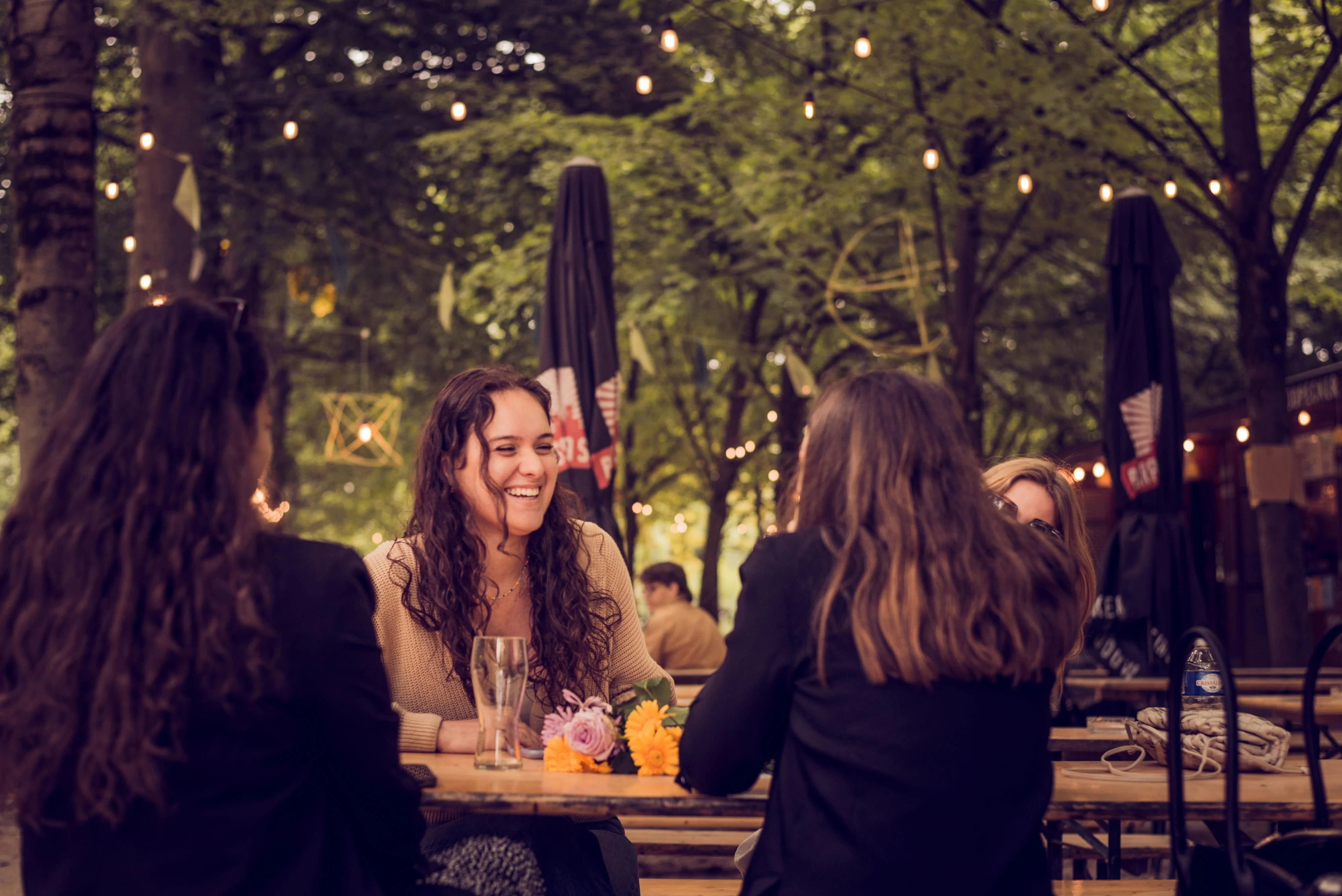 A group of friends sitting at an outdoor café table, smiling and engaging in conversation. The setting is adorned with string lights and trees in the background, creating a warm and inviting atmosphere.