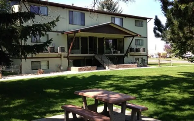 Front exterior of Twin Cedars Apartments in Helena, Montana, with large green lawn, mature trees, and picnic table under shaded area