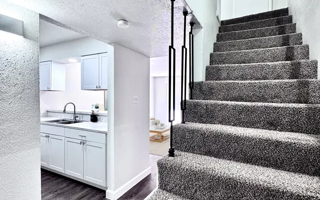 Modern Interior Staircase A well-lit staircase leading to an upper level, with carpeted steps and a modern railing. The upper area is seen invitingly open with a door at the top. The left side shows a glimpse of a kitchen area with white cabinetry and a modern faucet.