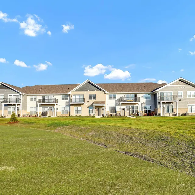 A modern apartment complex set against a clear blue sky with scattered clouds. The building features multiple units with balconies and large grassy areas in the foreground.