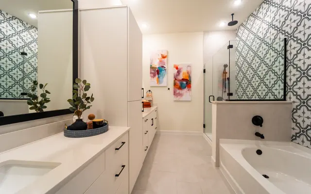 A modern bathroom featuring a white vanity with black accents, a large mirror, and decorative wall art. The walls are adorned with a patterned tile design, and there is a glass shower enclosure alongside a stylish bathtub.