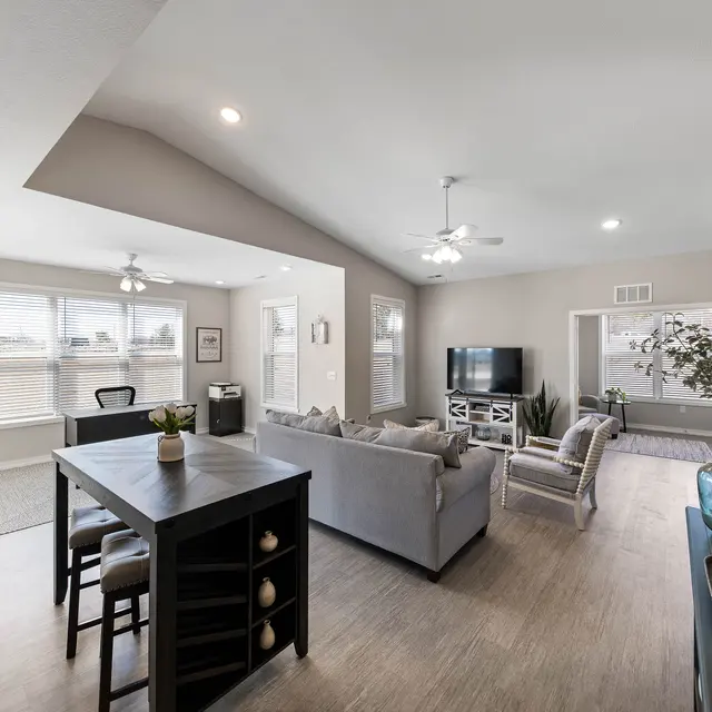 The Links at Franklin Apartments for Rent in Franklin IN - The Links at Franklin - Living Room with Wood-Style Plank Flooring, Table with Chairs, Grey Couch, TV Stand with TV, Armchair, Office Area, Mirror, House Plants, Large Windows with Blinds, and Ceiling Fan