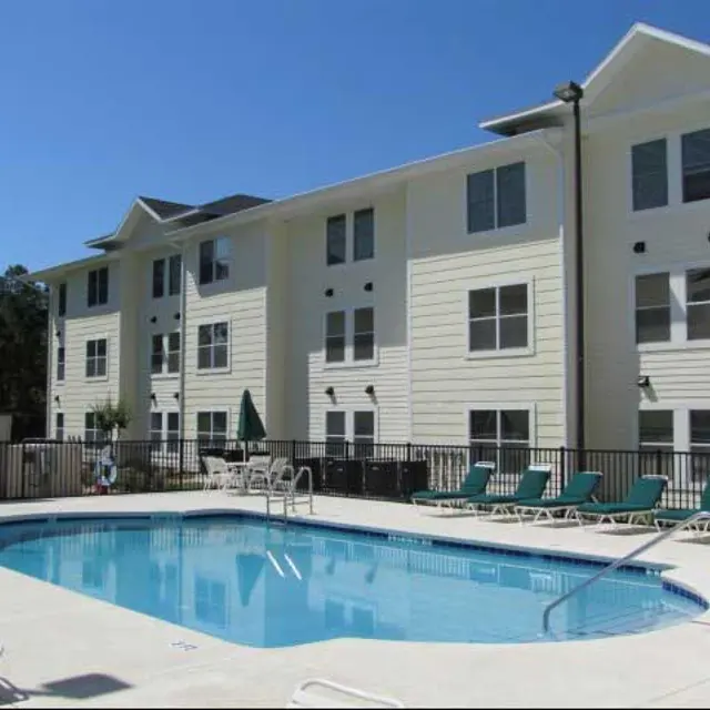 A pool area adjacent to a multi-story apartment building with lounge chairs and a sunny sky.