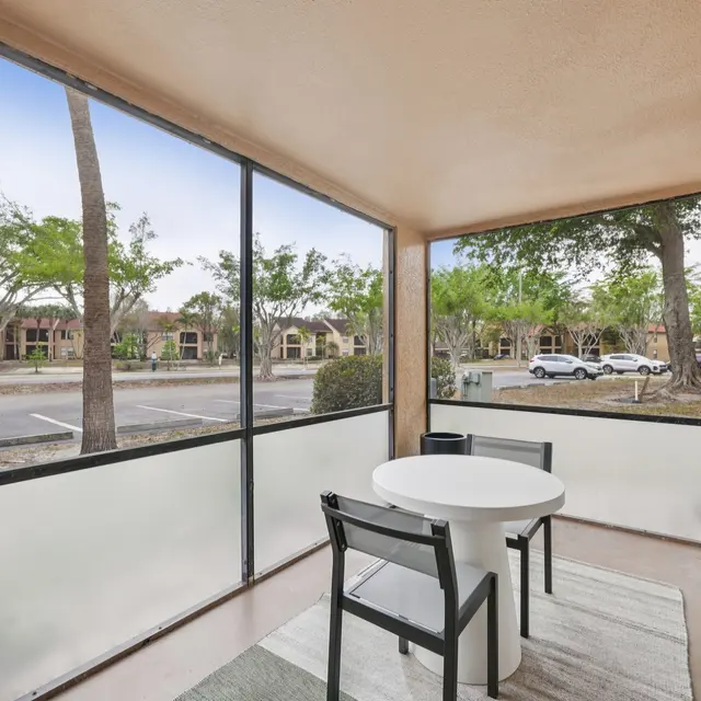 A screened patio area featuring a round white table and two black chairs, with a view of trees and a parking lot framed by modest buildings in the distance.