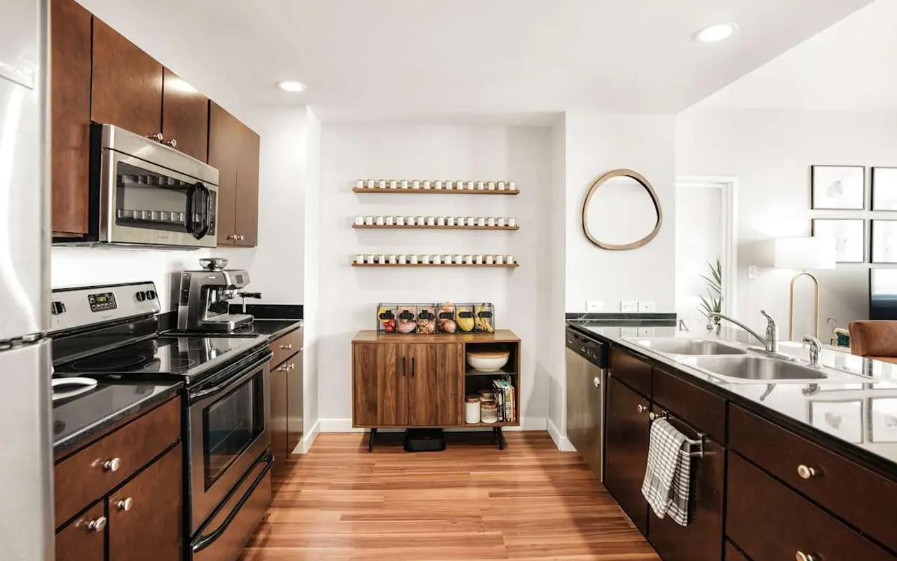 A modern kitchen with dark wooden cabinets, stainless steel appliances, and a large countertop. A wooden shelf with jars is mounted on the wall, and a wooden storage unit sits below. The flooring is wooden, and the space is well-lit with overhead lights.