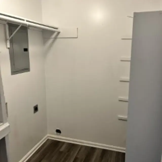 A small, empty room with a wooden floor, white walls, and a closet. The room has a door on the right and a built-in shelf on the back wall. A metal panel is mounted on the left wall.