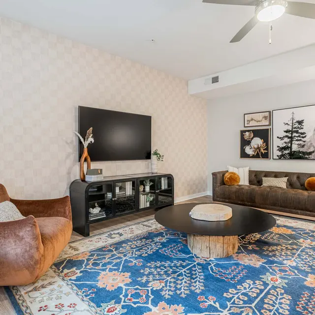 A stylish living room featuring a large flat-screen TV mounted on the wall, a dark sofa adorned with colorful pillows, a round coffee table, and a plush armchair. The floor is covered with a decorative area rug, and the walls have a light patterned wallpaper. A plant adds a touch of greenery to the space.
