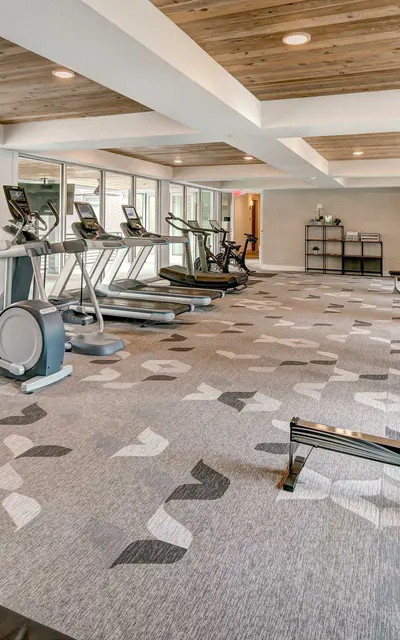 Fitness center area of our apartment community furnished with various exercise equipment, carpet flooring, and a high ceiling at 120 Ninth Street Apartments in San Antonio.