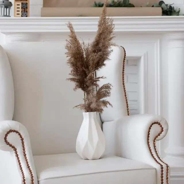 A cozy white armchair with decorative nailhead trim, featuring a vase of pampas grass on a light-colored seat in a home setting.