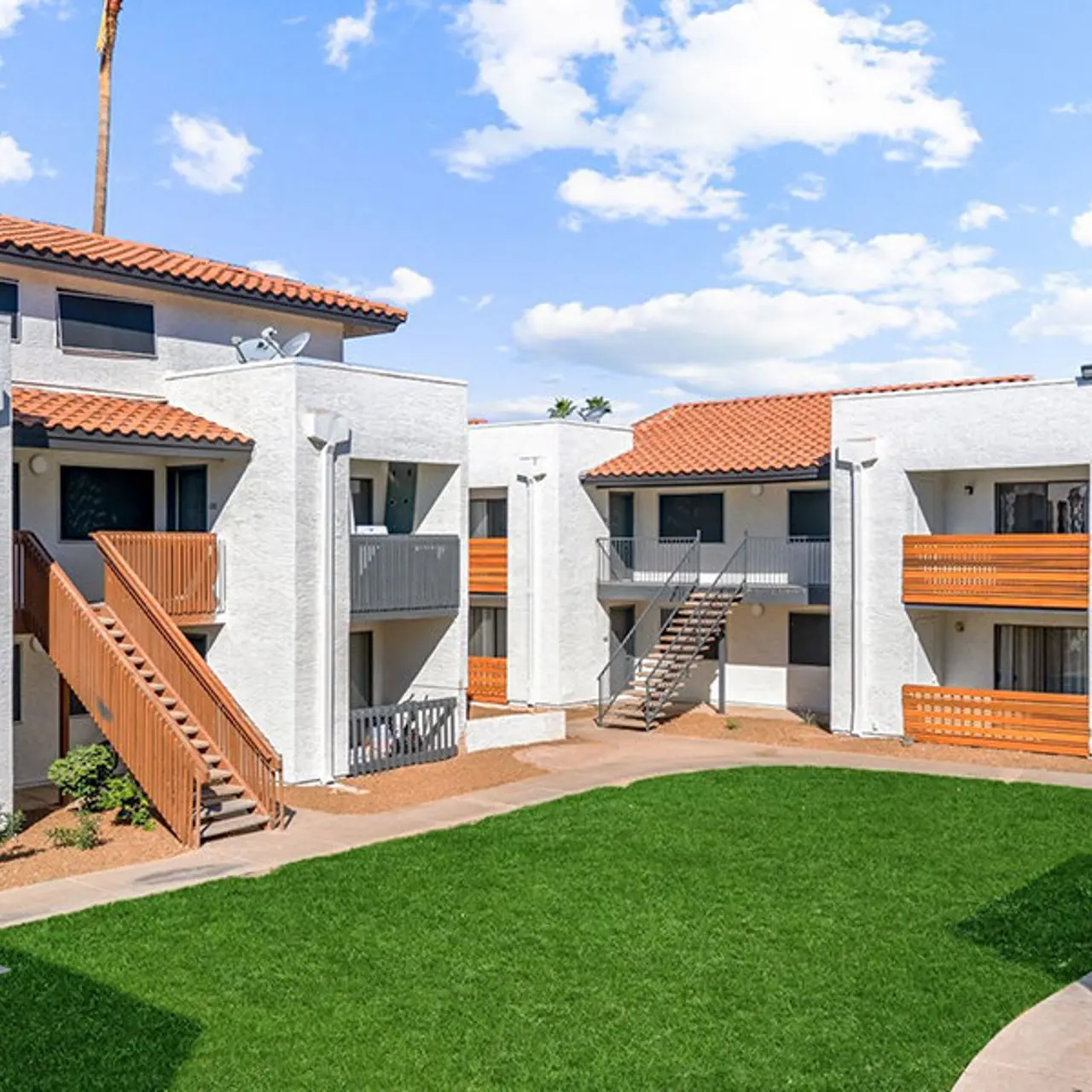 A view of an apartment complex with two buildings, featuring multiple balconies and stairs, surrounded by a lush green lawn and clear blue skies.