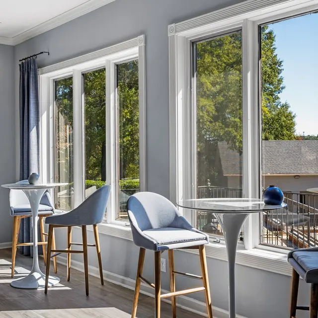 A modern seating area with gray walls and large windows allowing natural light. There are two small round tables and four stylish chairs. The area features greenery outside the window, creating a cozy atmosphere.