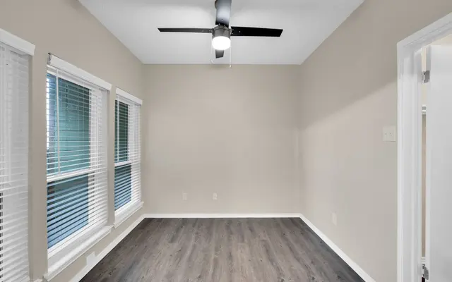 An empty room featuring a ceiling fan, large windows with blinds, and a dark wood floor.