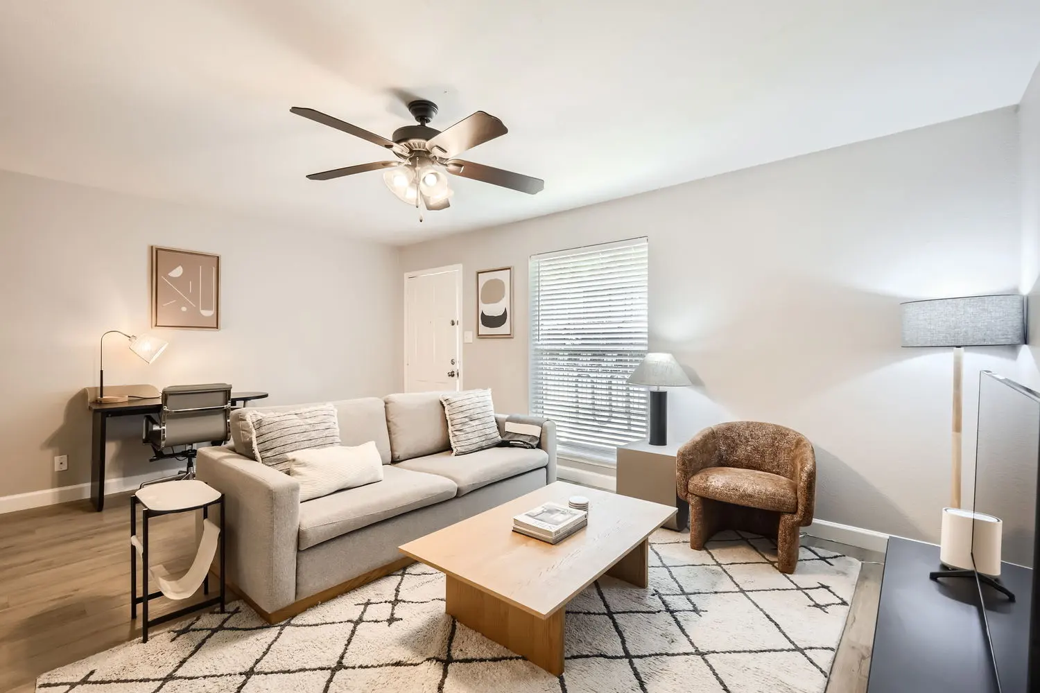 A cozy living room featuring a light gray sofa with cushions, a wooden coffee table, a small armchair, a desk with a lamp, and a television stand. The room has natural light from a window, a ceiling fan, and a patterned rug on the floor.