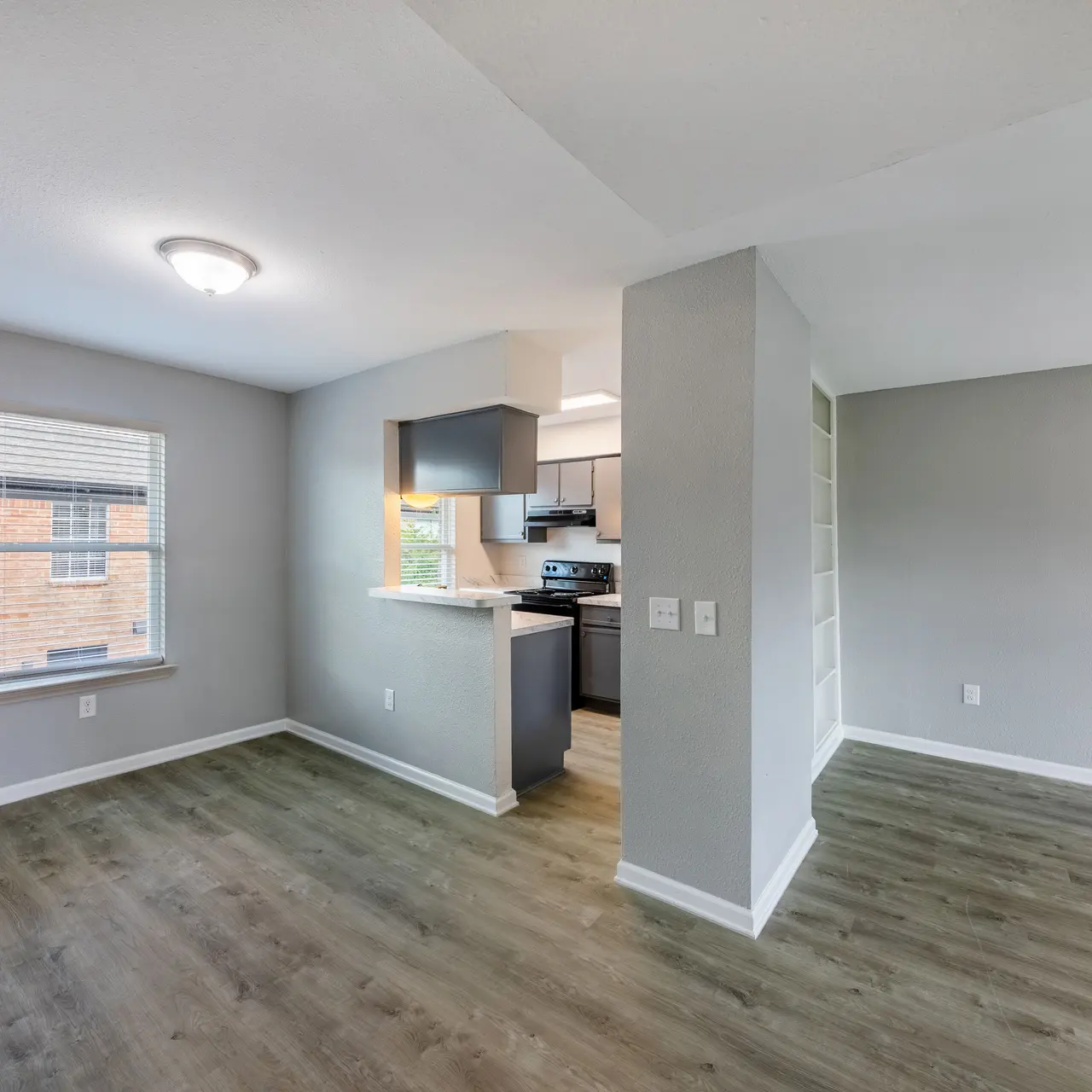 A modern apartment interior with an open floor plan featuring a living area with hardwood floors, a small kitchen bar, and a view of the kitchen appliances. The walls are painted light gray and there is plenty of natural light from the window.