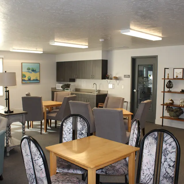 A modern living area with a kitchen, dining tables, and comfortable seating. The space features light-colored walls, wooden furniture, and decorative shelving units.