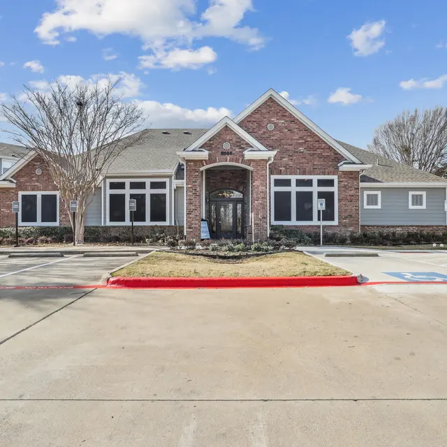 A brick building with large windows and a gabled roof, surrounded by a parking area and trees.