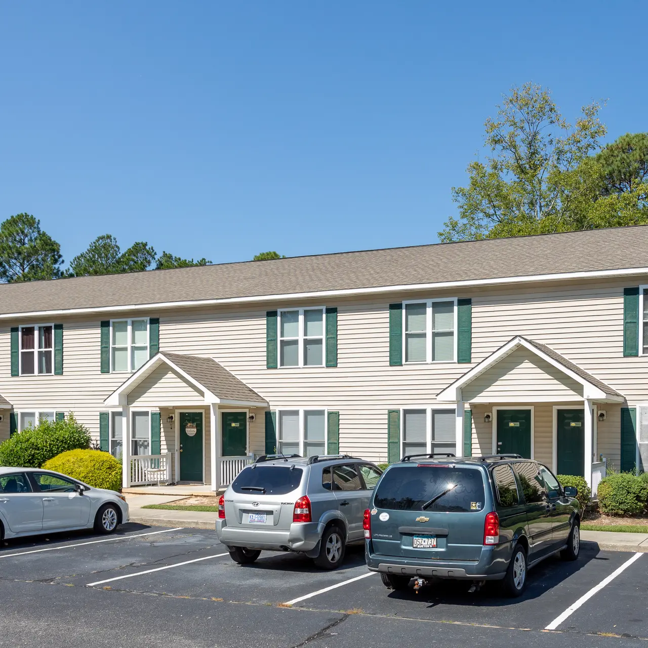 A two-story apartment complex with several parking spaces in front. The building has beige siding and green shutters, with bright sunlight illuminating the scene. There are neatly trimmed bushes and trees in the background.