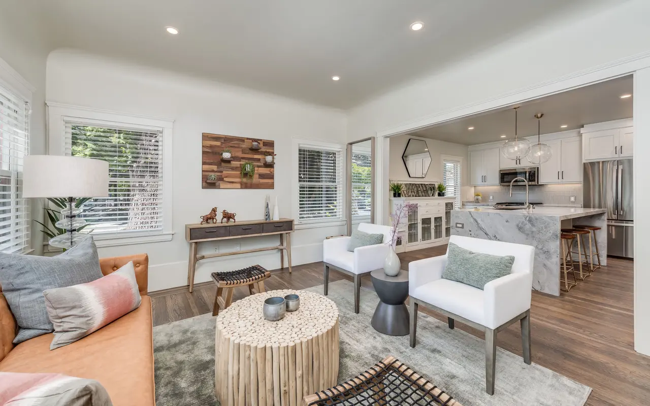A modern living room featuring an orange couch, white chairs, a round coffee table made of natural materials, and a stylish light fixture. The room is bright with large windows and an open view into the kitchen area, which includes gray countertops and contemporary appliances.