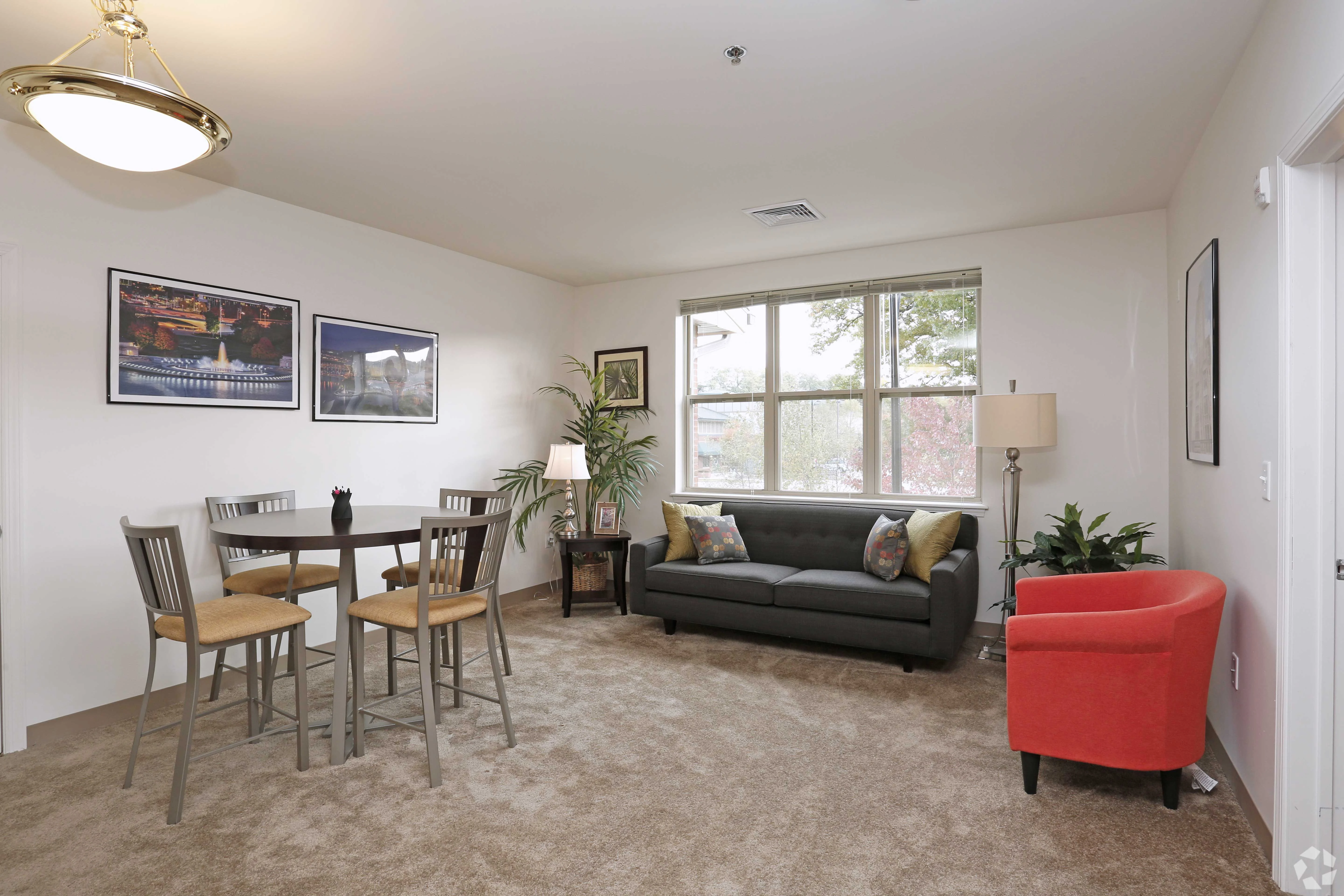 Cozy Living Room and Dining Area A cozy living room with a dining area, featuring a round table and four chairs, a gray sofa with colorful cushions, a red accent chair, and plants by the windows.