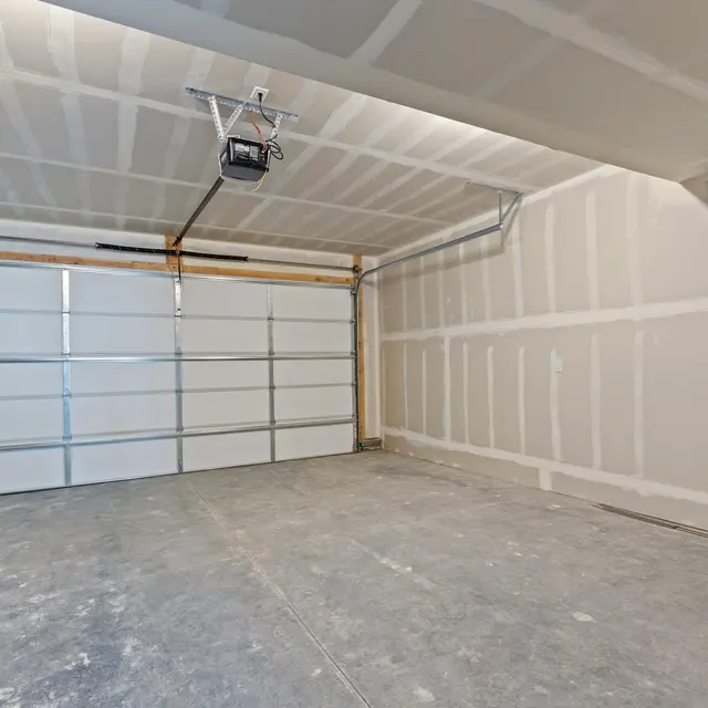A spacious, empty garage with bare drywall and a concrete floor. The garage door is closed, and there is a garage door opener mounted on the ceiling.